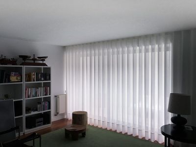 Cortina de voile branca a cobrir uma parede envidraçada numa sala de estar com estante e tapete verde