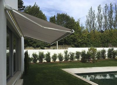 Toldo extensível bege sobre janela de casa branca, com jardim, sebe e piscina ao fundo