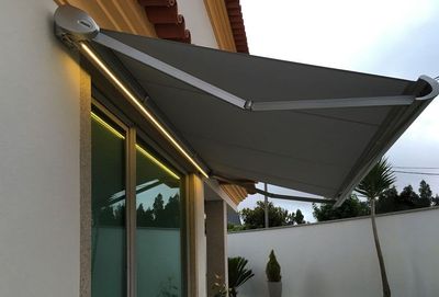 Toldo extensível cinzento escuro com fita LED acesa sobre janela de moradia ao entardecer