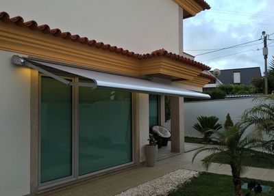 Toldo extensível bege sobre porta envidraçada de moradia com palmeira e jardim ao entardecer