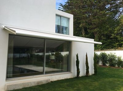 Toldo extensível branco sobre uma grande janela de moradia moderna branca, com relvado e ciprestes