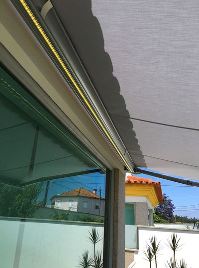 Detalhe da barra frontal de um toldo cinzento com fita LED sobre varanda envidraçada