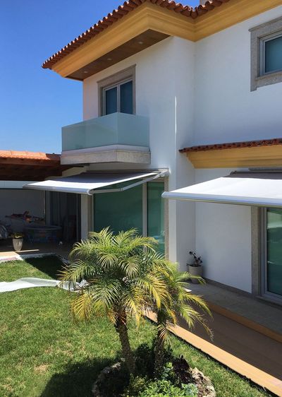 Dois toldos extensíveis brancos abertos sobre janelas de moradia com palmeira e céu azul