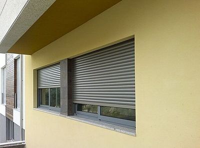 Estore exterior de réguas cinzentas semiaberto numa janela de moradia com fachada amarela