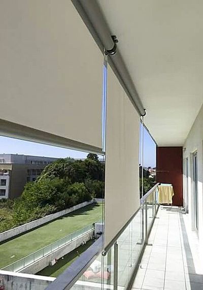 Estores exteriores de rolo bege numa varanda de apartamento com guarda de vidro e vista de jardim