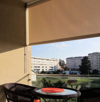 Estore exterior de rolo bege em varanda com mesa e cadeiras de vime e vista urbana ao entardecer