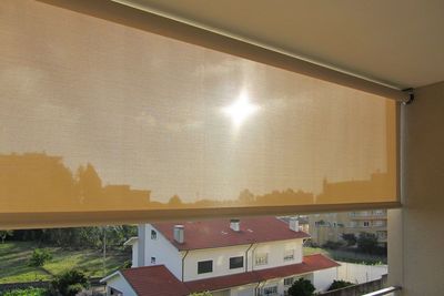 Estore exterior de rolo translúcido com o sol a atravessar o tecido, numa varanda sobre moradias