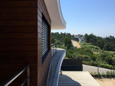 Fachada revestida a madeira com estore exterior de lâminas e varanda curva sobre paisagem verde