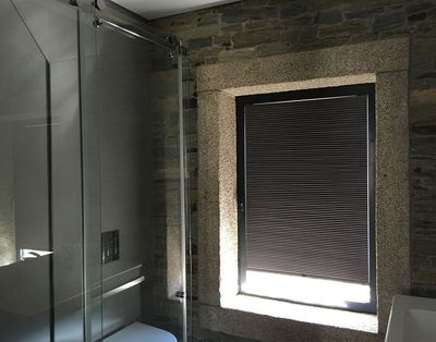 Estore de rolo numa janela com moldura de granito, numa casa de banho em pedra com duche