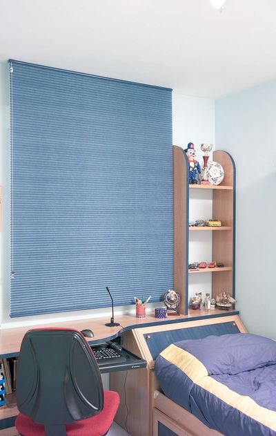 Estore de rolo em tecido azul numa janela de quarto juvenil com secretária, estante e cama