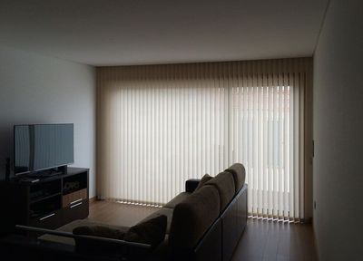 Cortina de lâminas verticais bege numa sala de estar com sofá castanho e móvel de TV