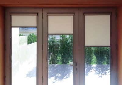Três estores de rolo bege a meia altura em portas envidraçadas com vista de jardim e sebe