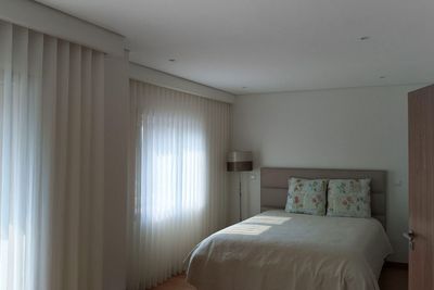 Cortinados de voile branco até ao chão num quarto com cama, almofadas florais e candeeiro de pé