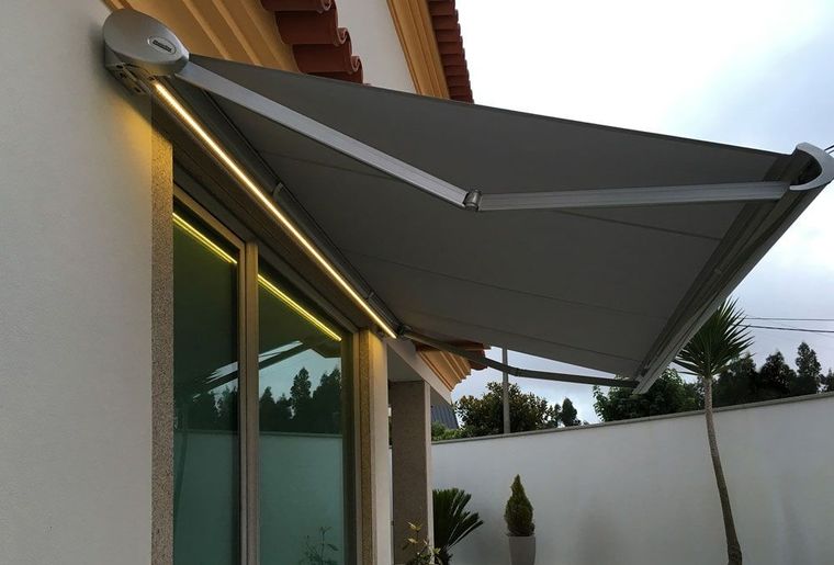 Toldo extensível cinzento escuro com fita LED acesa sobre janela de moradia ao entardecer
