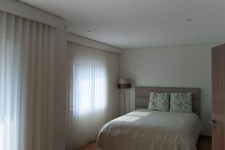 Cortinados de voile branco até ao chão num quarto com cama, almofadas florais e candeeiro de pé